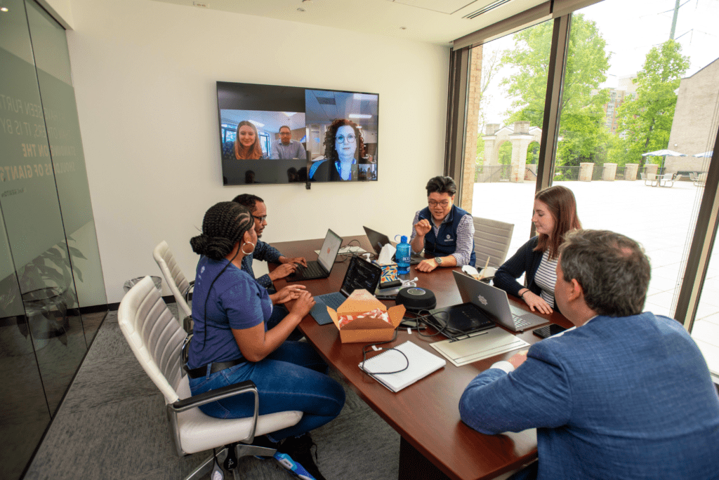 A group photo of MetroStar employees working in a conference room with two employees dialing in remotely